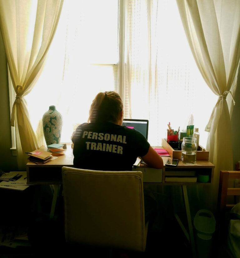 A woman in a "Personal Trainer" shirt sits at a sunlit desk, working at her computer with her back to the camera.