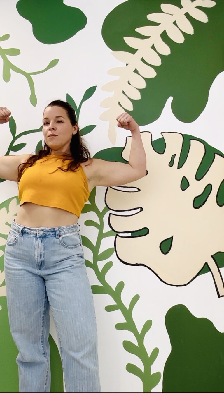 Gwen, FIT by GP coach, flexing both arms in front of a green botanical mural — strength training for women 40+