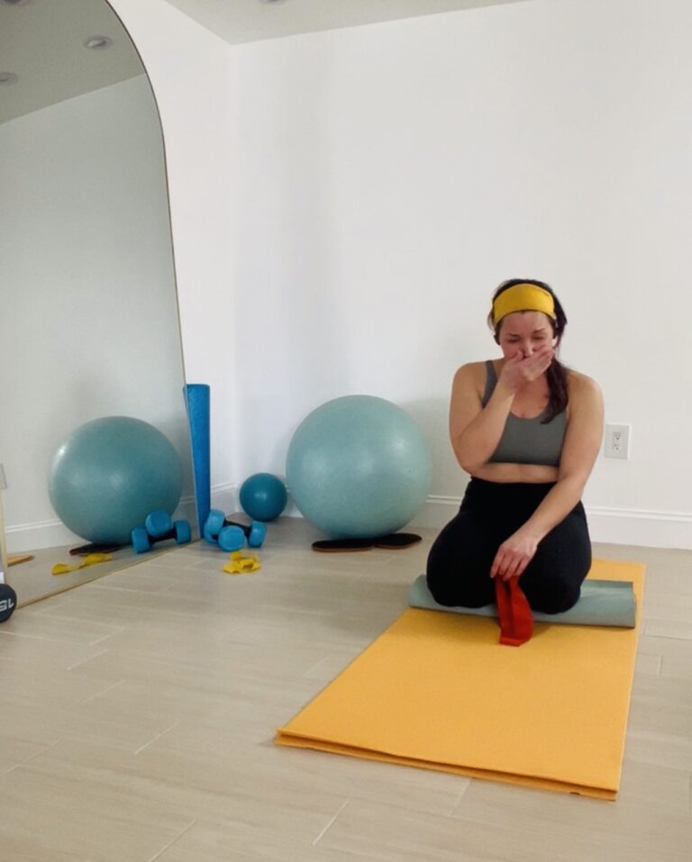 Gwen kneeling on an orange mat in her home training space, holding a resistance band, breaking down in tears mid-workout. Dumbbells, stability balls, and a foam roller around her.